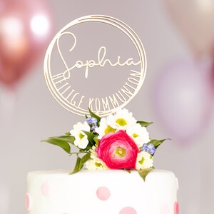 Può includere: Un topper per torta dorato con il nome "Sophia" e il testo "Heilige Kommunion" in un cerchio. Il topper è su una torta bianca con pois rosa e un bouquet di fiori bianchi e rosa.