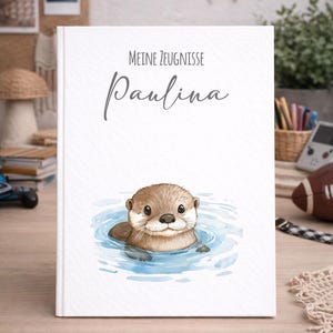 Può includere: Un libro bianco con il testo "Meine Zeugnisse Paulina" e un'illustrazione ad acquerello di una lontra in acqua blu. La lontra ha la pelliccia marrone e guarda l'osservatore. Il libro è su un tavolo di legno.