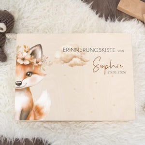 Op de afbeelding: Houten herinneringskist met een illustratie van een vos met een bloemenkrans. De kist is gepersonaliseerd met de naam "Sophie" en de datum 23.01.2026. Een kleine teddybeer en houten blokken zijn ook zichtbaar.