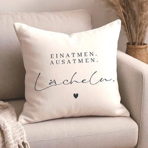 Può includere: Cuscino quadrato color crema con testo nero "EINATMEN. AUSATMEN. Lächeln." e un piccolo cuore nero. Il cuscino è su un divano beige, con una coperta beige e pampas sullo sfondo.