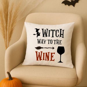 Può includere: Cuscino quadrato bianco sporco con la scritta "Witch Way to the Wine" in nero e rosso. Sono presenti anche una strega su una scopa, una scopa e un calice di vino. Il cuscino è su una sedia beige con una zucca in primo piano.