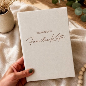 Könnte beinhalten: Ein rechteckiges Buch mit einem hellbeigen Stoffeinband. Auf dem Vorderdeckel steht das Wort STAMMBUCH in einer einfachen serifenlosen Schrift über dem Namen Familie Kath, der in einer dunkelbraunen Schreibschrift gedruckt ist.