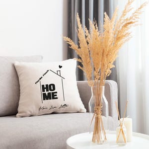 Può includere: Un cuscino bianco con testo nero che dice "HOME" con le parole "Forte Mack" e "Kai Lia Letta" sopra e sotto la parola "HOME". Il cuscino è su un divano grigio chiaro con un vaso di erba della pampa secca su un tavolo bianco davanti al divano.