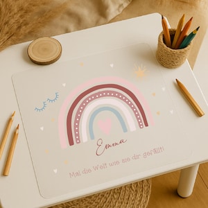 Könnte beinhalten: Eine hellbeige Tischunterlage mit einem Regenbogen-Design in Rosa, Burgunder und Blau, einem Herz und dem Namen "Emma". Der Text "Mal die Welt wie sie dir gefällt!" ist ebenfalls vorhanden. Ein kleiner Korb mit Bleistiften und ein Holzstift liegen auf dem Tisch.