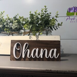 May include: A wooden sign with the word "Ohana" written in white paint. The sign is sitting on a white shelf with a vase of greenery behind it.