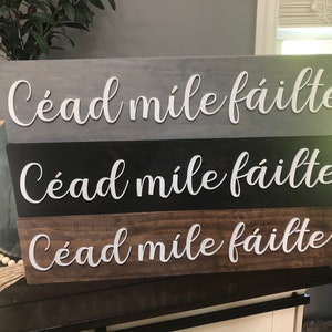 May include: Three wooden signs with the text "Cead mile failte" in white lettering. The signs are stacked on top of each other, with the top sign being gray, the middle sign being black, and the bottom sign being brown.