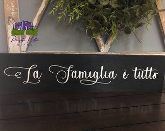 May include: Black wooden sign with white cursive lettering that reads "La Famiglia è tutto".