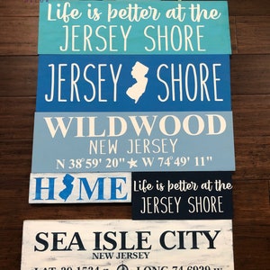 May include: Five wooden signs with blue and white paint, featuring the text "Life is better at the Jersey Shore", "Jersey Shore", "Wildwood New Jersey", "Home", and "Sea Isle City New Jersey". The signs also include coordinates for Wildwood and Sea Isle City.