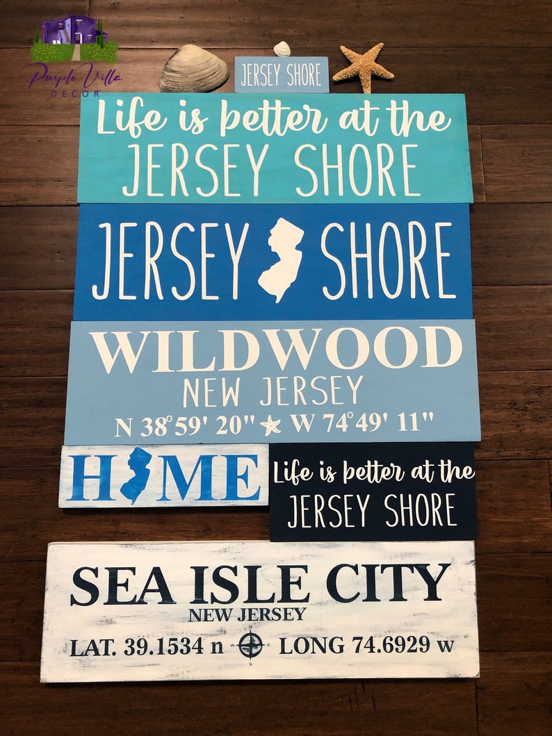 May include: Five wooden signs with blue and white paint, featuring the text "Life is better at the Jersey Shore", "Jersey Shore", "Wildwood New Jersey", "Home", and "Sea Isle City New Jersey". The signs also include coordinates for Wildwood and Sea Isle City.