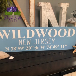 May include: A light blue wooden sign with white lettering that reads "WILDWOOD NEW JERSEY N 38&deg;59' 20" W 74&deg;49' 11"".