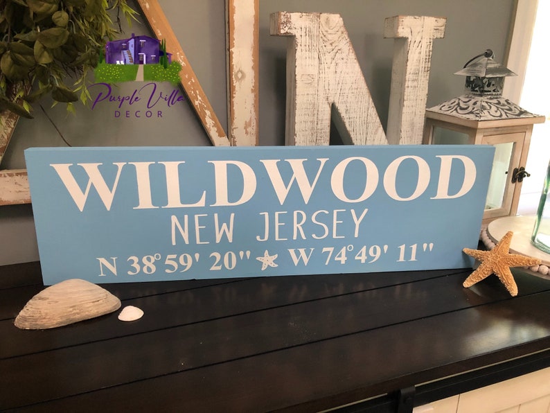 May include: A light blue wooden sign with white lettering that reads "WILDWOOD NEW JERSEY N 38&deg;59' 20" W 74&deg;49' 11"".