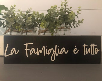 May include: A black wooden sign with white lettering that reads "La Famiglia è tutto". The sign is sitting on a table with three glass jars filled with greenery.