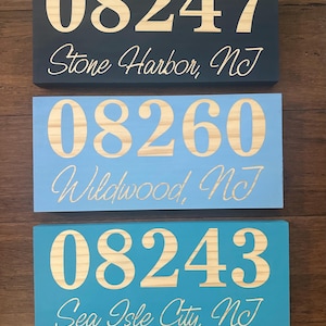 May include: Three wooden signs with white numbers and text. The top sign is black with the numbers 08247 and the text "Stone Harbor, NJ". The middle sign is light blue with the numbers 08260 and the text "Wildwood, NJ". The bottom sign is teal with the numbers 08243 and the text "Sea Isle City, NJ".
