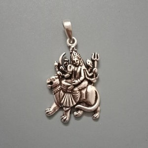 Może przedstawiać: Srebrny wisiorek przedstawiający szczegółową scenę z hinduską boginią Durgi jadącą na lwie. Jest przedstawiona z wieloma ramionami trzymającymi różne bronie, w tym trójząb i miecz.