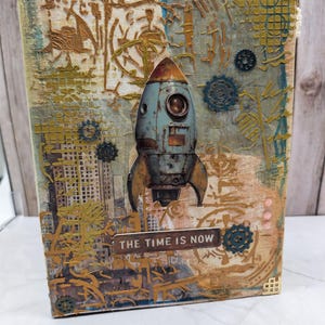 Pode incluir: Uma obra de arte em técnica mista com um foguete em estilo vintage contra um fundo texturizado com engrenagens e elementos arquitetônicos. A frase "THE TIME IS NOW" é exibida em uma pequena placa. A paleta de cores inclui tons de azul, dourado e marrom.