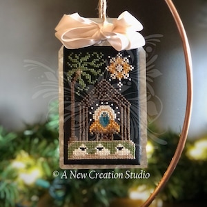 Può includere: Un ornamento natalizio a punto croce raffigurante una scena della Natività su sfondo nero. L'ornamento è adornato da un nastro beige e sospeso da uno spago. Il disegno comprende una stella, palme e una stalla. Il testo "A New Creation Studio" è in basso.