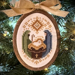 Könnte beinhalten: Ein ovales Kreuzstich-Ornament mit einer Krippenszene, bei der Figuren in grünen und blauen Gewändern eine zentrale Krippe umgeben. Das Design umfasst einen goldenen Stern darüber und eine goldene Schleife in einem braunen Holzrahmen.