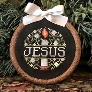 May include: A cross-stitch embroidery featuring the word "JESUS" in white, surrounded by a candle and floral design on a black background. The embroidery is set in a round, wooden frame and adorned with a white satin bow at the top. The overall theme is religious.