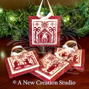 May include: Four red and white Christmas ornaments with a nativity scene design. The ornaments are made of fabric and have a white background with red stitching. The ornaments are hanging from a white ribbon.