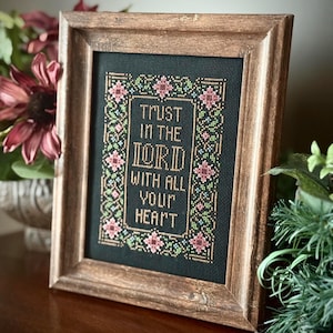 May include: A framed cross-stitch with the text "TRUST IN THE LORD WITH ALL YOUR HEART." The embroidery features a floral border in pink, green, and gold against a black background. The frame is made of wood with a warm, brown finish.