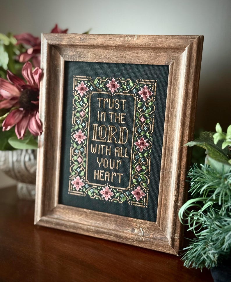 May include: A framed cross-stitch with the text "TRUST IN THE LORD WITH ALL YOUR HEART." The embroidery features a floral border in pink, green, and gold against a black background. The frame is made of wood with a warm, brown finish.