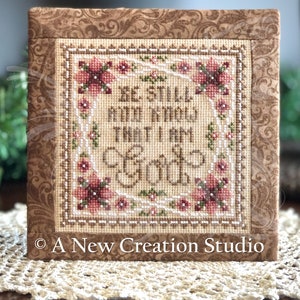 May include: A framed cross-stitch piece with the text "BE STILL AND KNOW THAT I AM GOD" surrounded by floral accents. The piece is in a square frame with a brown floral pattern and a beige background. The words "A New Creation Studio" are at the bottom.