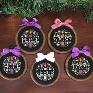 Könnte beinhalten: Fünf bestickte Weihnachtsschmuckstücke mit Holzreifen und schwarzem Stoff. Jedes zeigt ein Kerzenmotiv und ein Wort: Frieden, Freude, Hoffnung, Jesus und Liebe. Die Ornamente sind mit bunten Schleifen verziert.