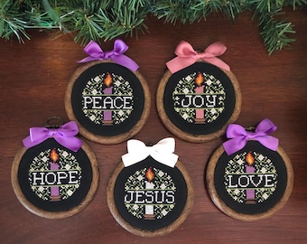 Op de afbeelding: Vijf geborduurde kerstversieringen met houten hoepels en zwarte stof. Elk heeft een kaarsontwerp en een woord: Vrede, Vreugde, Hoop, Jezus en Liefde. De ornamenten zijn versierd met kleurrijke strikken.
