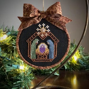 Può includere: Un ornamento circolare a punto croce con sfondo in tessuto nero e una scena della natività ricamata. Il design include una stalla marrone, una figura verde, una blu, una forma centrale viola e un fiocco marrone con motivo decorativo.
