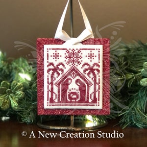 May include: A red and white Christmas ornament with a cross-stitch design of the nativity scene. The ornament is hanging from a white ribbon and is on a gold stand.