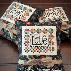 May include: Three square fabric pin cushions featuring black floral fabric borders and a central cream-colored cross-stitch panel. Each cushion displays a word in black thread: Faith, Hope, or Love, surrounded by a colorful geometric border and a burlap ribbon.