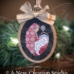 Peut inclure: Ornement ovale en point de croix avec un nœud en toile de jute et une bordure en ficelle. Le motif présente un motif en forme de cœur en rouge et blanc, et une figurine en bleu et blanc. Le texte "A New Creation Studio" est en bas.