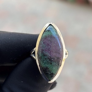 May include: A gold-toned ring featuring a large, oval-shaped stone with a mix of colors. The stone has a deep purple center, surrounded by green and red hues. The ring's band is a simple, polished gold.