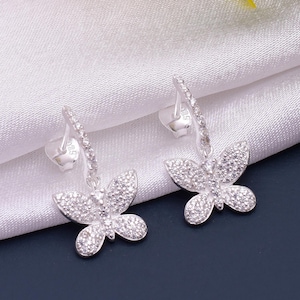 May include: A pair of silver butterfly earrings. Each earring has a butterfly pendant adorned with small, sparkling stones. The earrings are displayed against a white fabric background with a dark blue surface.