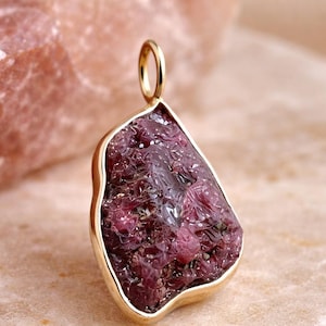 Puede incluir: Colgante dorado con una gema de rubí en bruto de forma irregular. El rubí tiene un tono rojo-púrpura intenso y una superficie texturizada. El colgante está engastado en un bisel dorado y suspendido de un pequeño lazo.