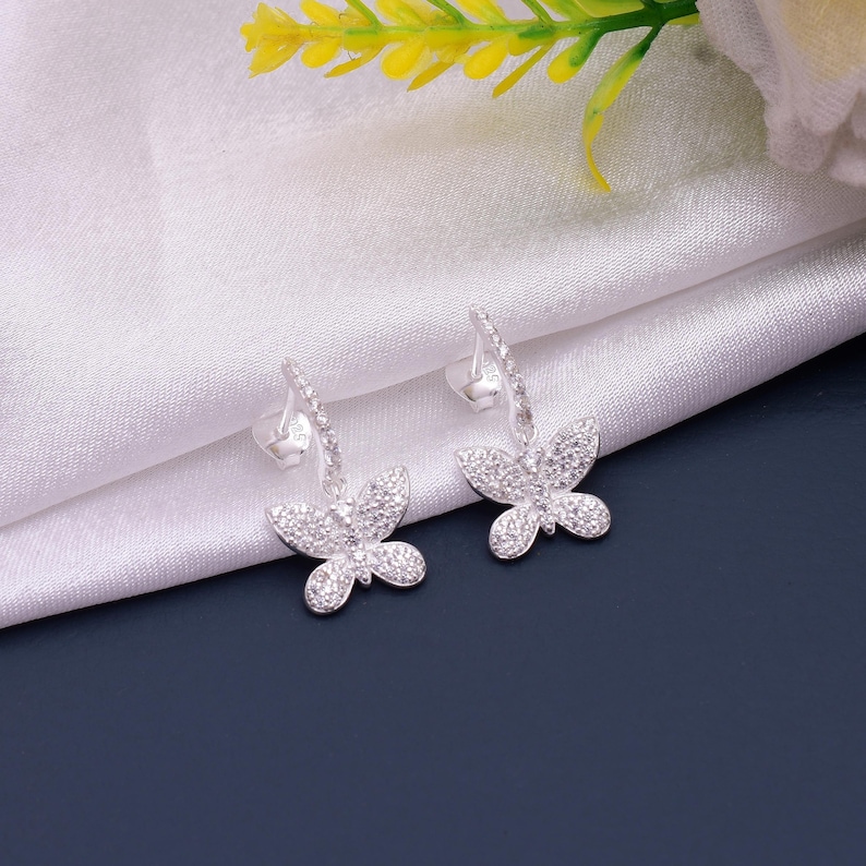 May include: A pair of silver butterfly earrings. Each earring has a butterfly pendant adorned with small, sparkling stones. The earrings are displayed against a white fabric background with a dark blue surface.