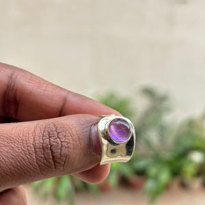 May include: A silver ring featuring a large, round, purple gemstone. The ring has a broad band and a simple, modern design. The gemstone is set in a bezel setting, and the ring is highly polished. A piece of jewellery.
