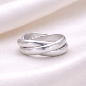 May include: A silver-colored ring with three intertwined bands. The bands are smooth and rounded, creating a continuous, interlocking design. The ring is set against a white, textured background, highlighting its metallic sheen.