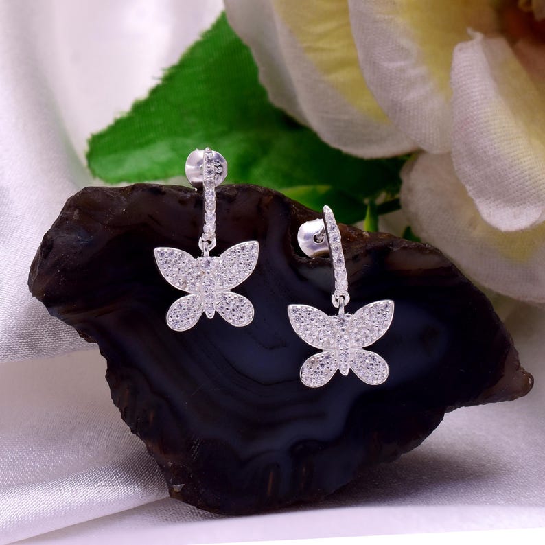 May include: Silver butterfly drop earrings. Each earring has a butterfly design, covered in small, sparkling stones. The earrings are shown on a dark, textured surface, with a white and green background.