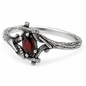 May include: A silver ring featuring a marquise-cut dark red garnet gemstone set within a band designed to resemble intertwined tree branches with small leaves. The metal has an oxidised finish that highlights the textured bark details.