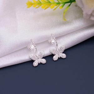 May include: A pair of silver butterfly earrings. Each earring features a butterfly pendant encrusted with small, sparkling stones. The earrings are set against a white fabric background with a dark blue surface.