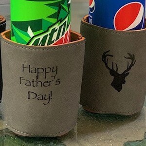 May include: Three different can coolers with personalized designs. The first cooler is black with a white graphic of a cartoon figure. The second cooler is gray with the text "Happy Father's Day!" The third cooler is gray with a black graphic of a deer head.