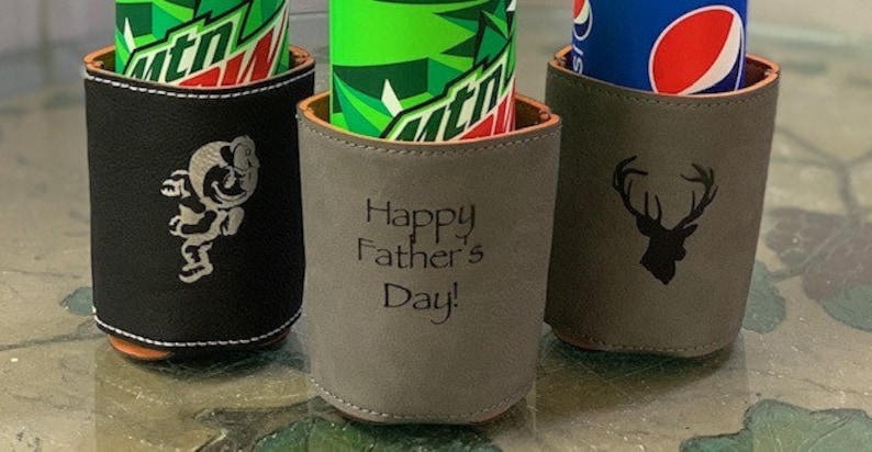 May include: Three different can coolers with personalized designs. The first cooler is black with a white graphic of a cartoon figure. The second cooler is gray with the text "Happy Father's Day!" The third cooler is gray with a black graphic of a deer head.