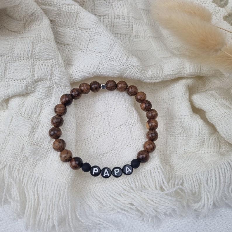 May include: A brown wooden beaded bracelet with black beads spelling out the word "PAPA".