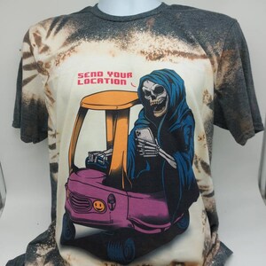 May include: A bleached grey t-shirt with a tie-dye pattern features a graphic of a skeleton wearing a blue hooded robe and driving a pink toy car. The text "SEND YOUR LOCATION" is printed above the graphic.