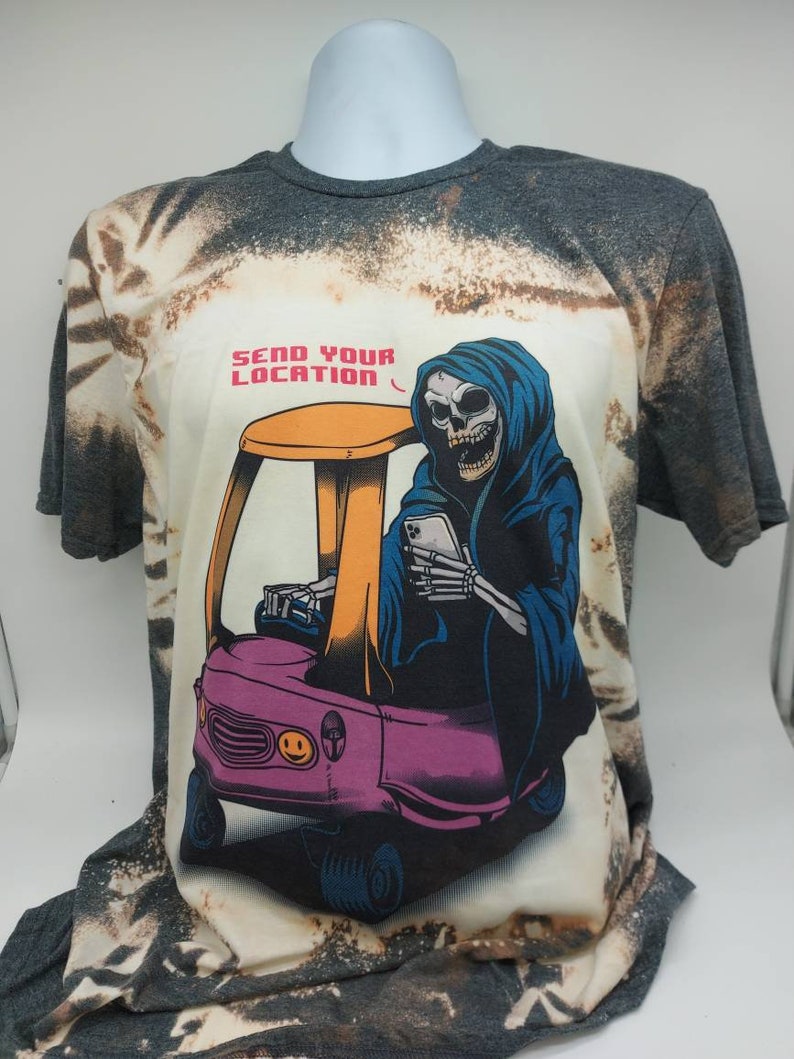May include: A bleached grey t-shirt with a tie-dye pattern features a graphic of a skeleton wearing a blue hooded robe and driving a pink toy car. The text "SEND YOUR LOCATION" is printed above the graphic.