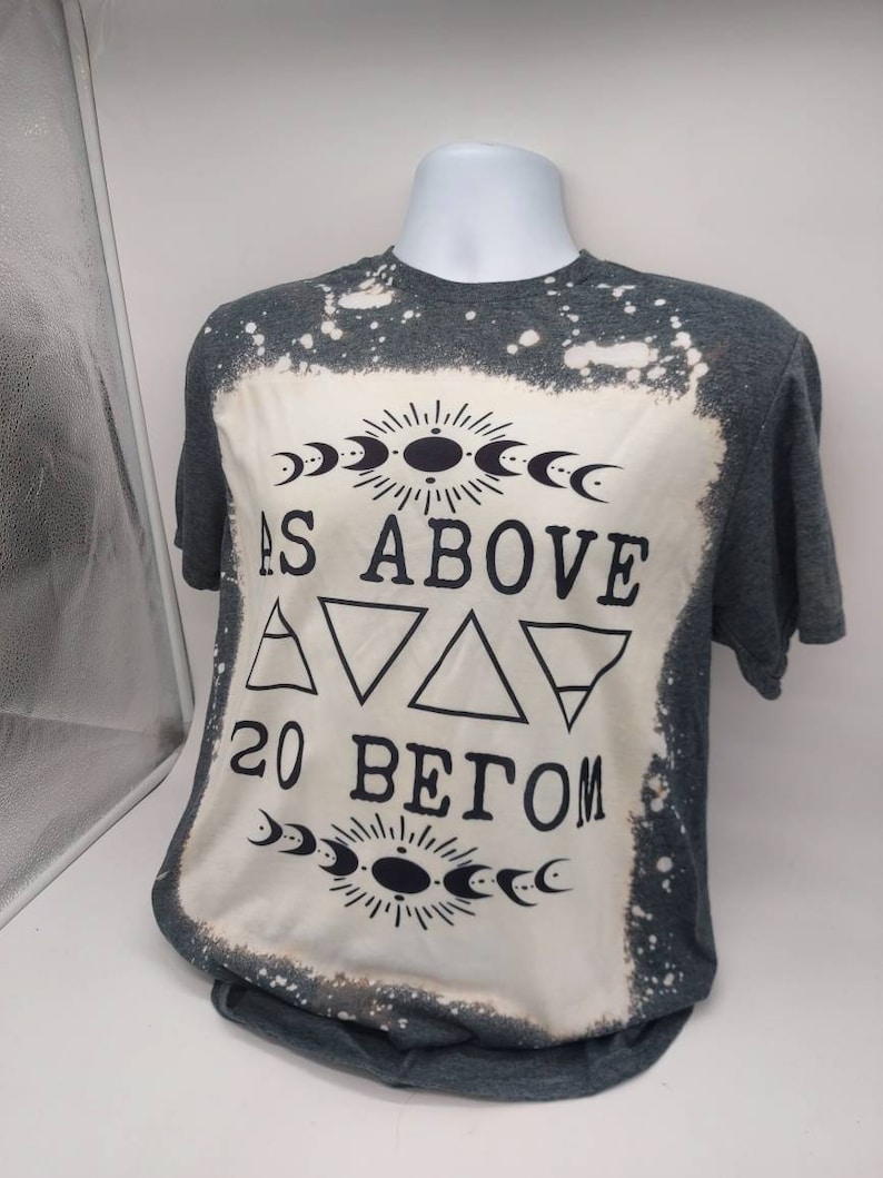 May include: A gray t-shirt with a bleached design featuring the text "As Above So Below" with a moon and sun symbol above and below the text. The design also includes three triangles and the number 20.
