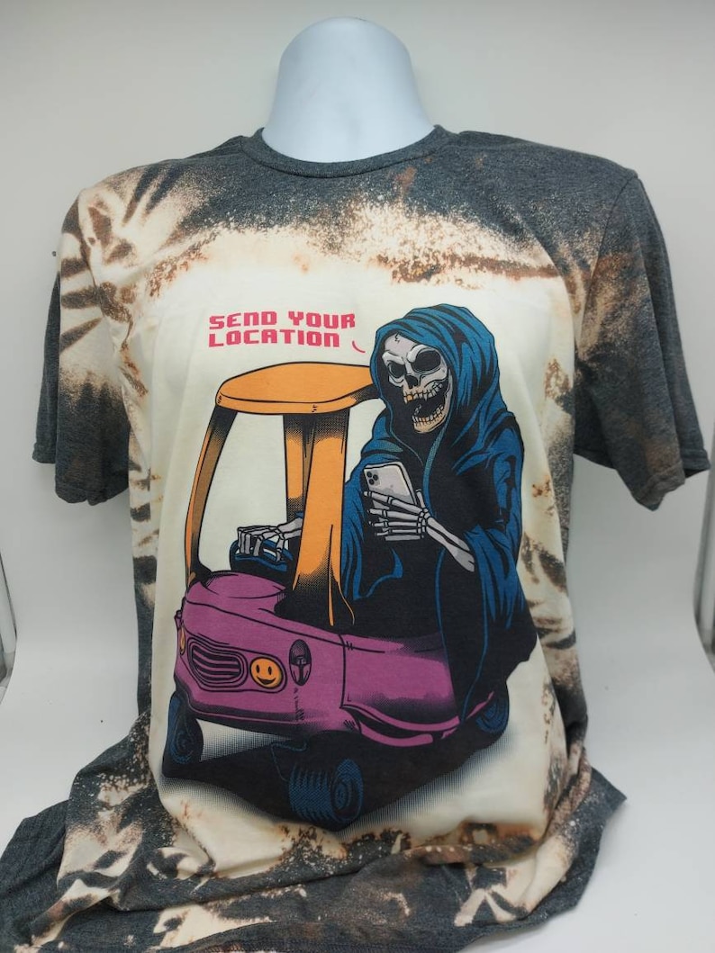 May include: A bleached grey t-shirt with a tie-dye pattern features a graphic of a skeleton wearing a blue hooded robe and driving a pink toy car. The text "SEND YOUR LOCATION" is printed above the graphic.