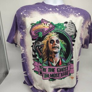 May include: A white t-shirt with a purple bleach design. The shirt features a graphic of a character from the movie Beetlejuice. The text on the shirt reads "You're the ghost with the most, babe." and "Never trust the living."