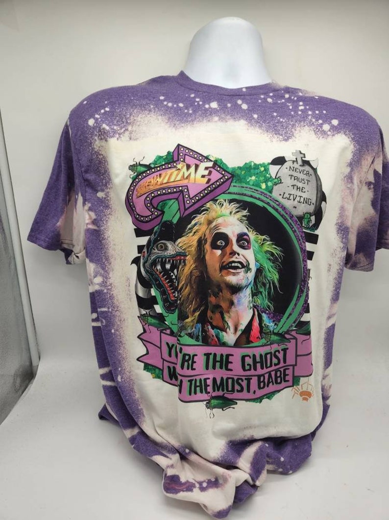 May include: A white t-shirt with a purple bleach design. The shirt features a graphic of a character from the movie Beetlejuice. The text on the shirt reads "You're the ghost with the most, babe." and "Never trust the living."